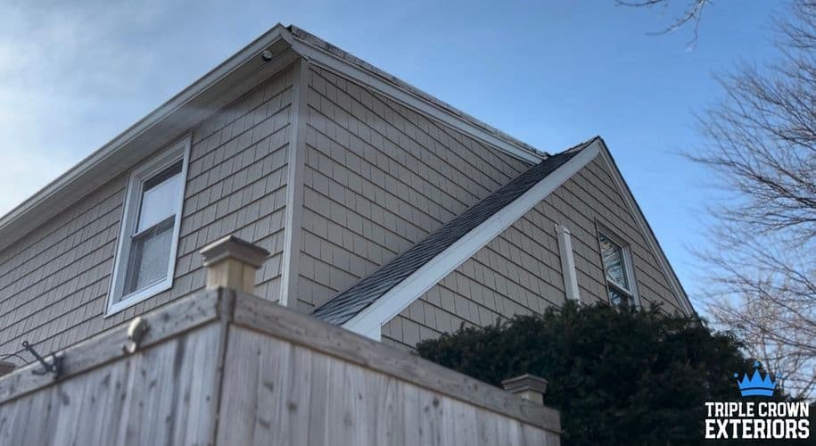 Exterior view of a house with siding and a fence against a clear sky. Triple Crown Exteriors logo.
