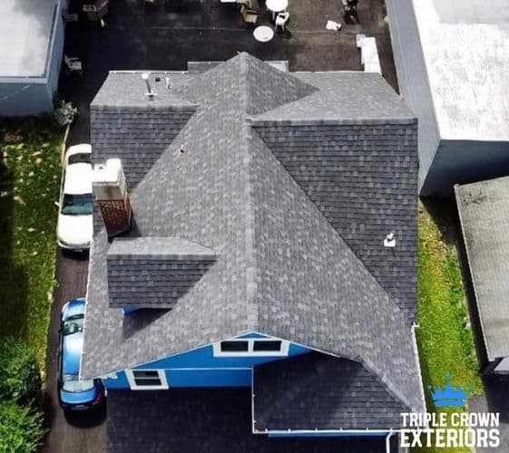 Aerial view of a blue house with a gray shingle roof, surrounded by greenery and parked cars.