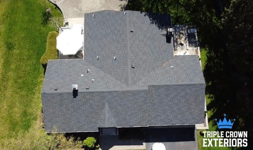 Aerial view of a large residential roof with dark shingles surrounded by green lawn.