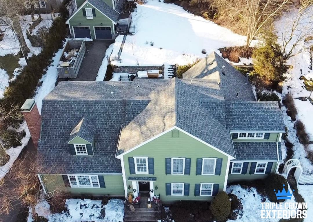 Aerial view of a green house with snow-covered yard and driveway, showcasing architectural details.
