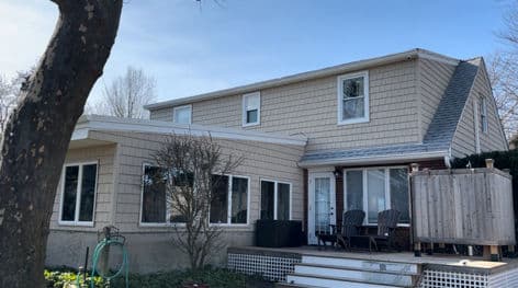 Two-story beige house with large windows, a porch, and outdoor seating in a suburban setting.
