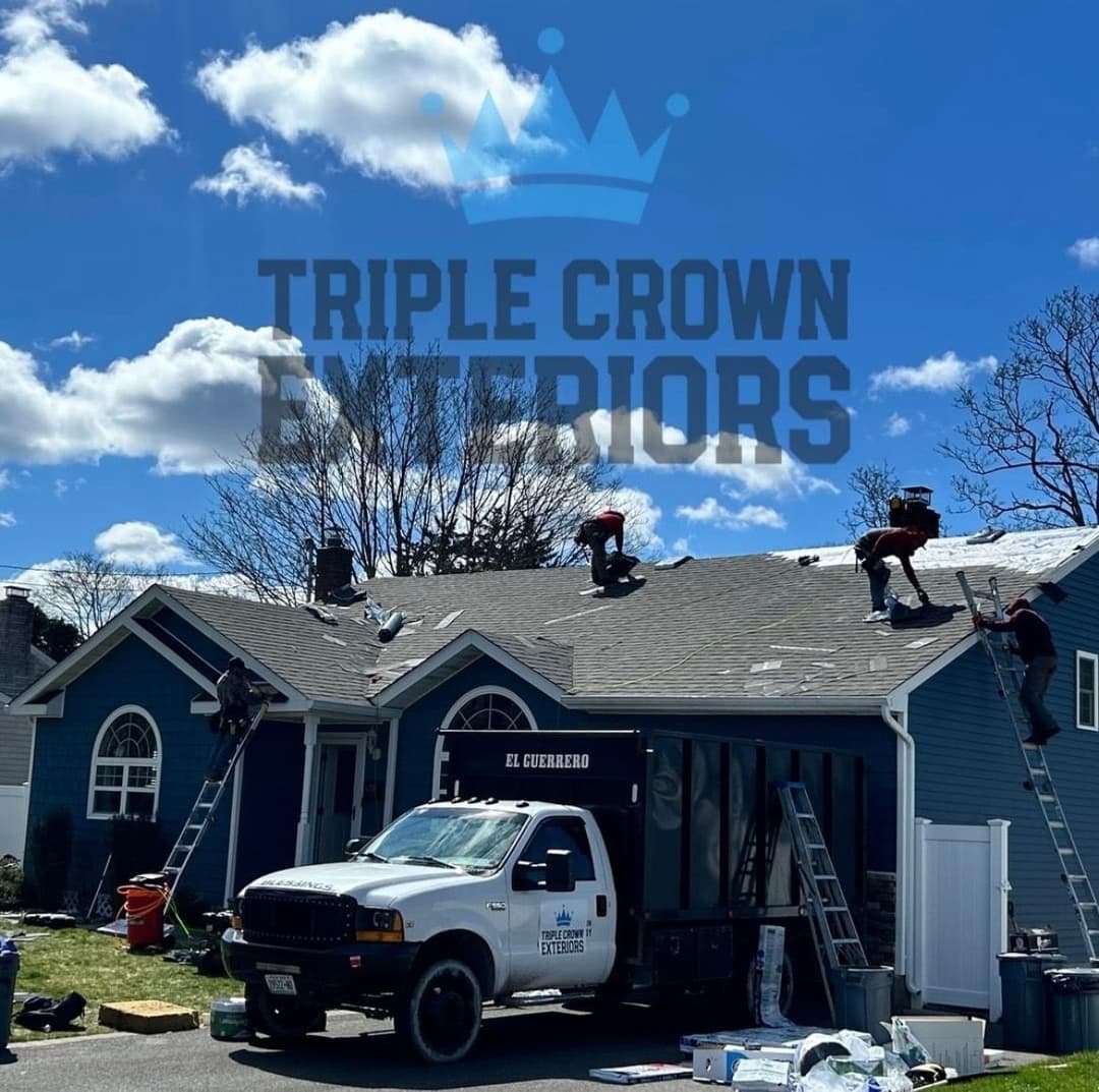 Roofing crew working on a home with Triple Crown Exteriors truck parked nearby under blue sky.