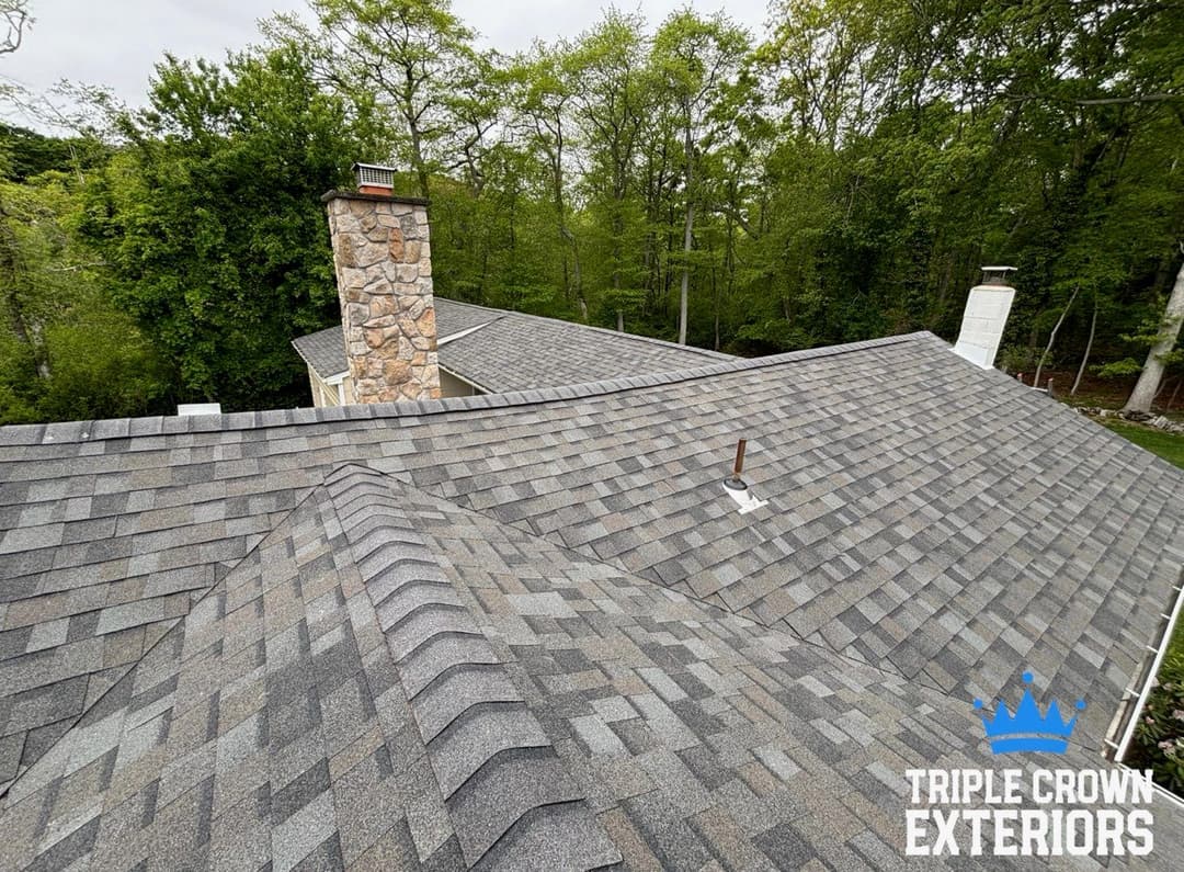 Gray shingle roof with chimney surrounded by greenery from a high viewpoint. Triple Crown Exteriors logo.